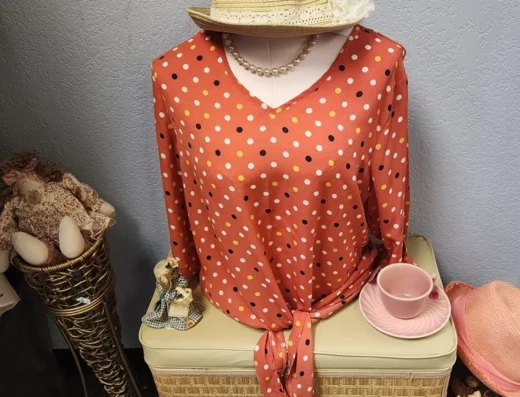 Mannequin wearing a coral polka-dot blouse with a knotted front, straw hat, and pearl necklace on a wicker trunk display.