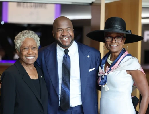 Three smiling adults posing together at a formal event; the man in the middle wears a navy suit and tie, flanked by an older woman in black and a woman in a wide-brim hat and pearl necklace on the right.