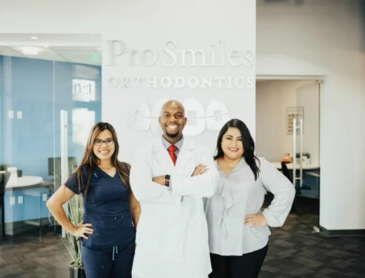 Three smiling dental professionals pose in the ProSmiles Orthodontics lobby with the logo behind them.