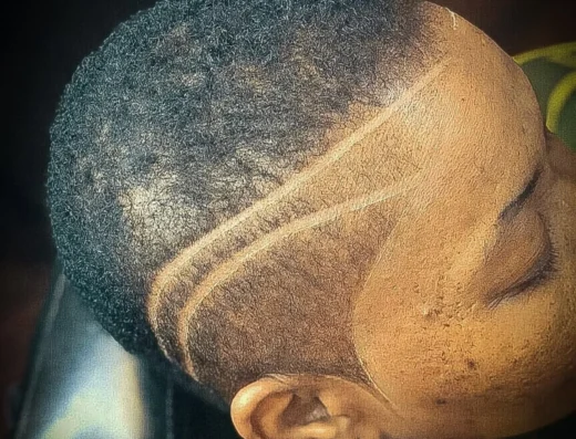 Close-up of a young person’s head showing a fresh fade with a bold curved line shaved into the side. The person wears a pacifier and a yellow clip visible near the ear.