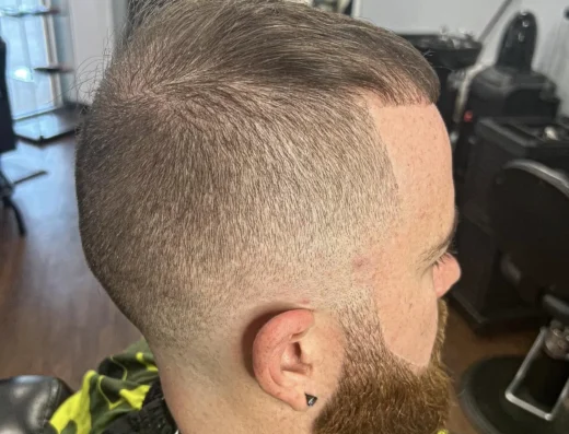 Side view of a man with a high skin fade on the sides, longer hair brushed over the top, and a full brown beard; he wears a yellow-and-black patterned barber cape in a barber shop.