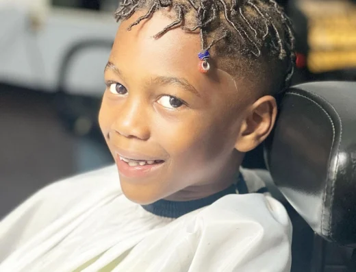 Smiling young boy with short twisted braids wearing a white barber cape inside a car or salon chair.