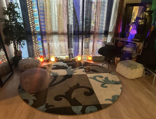Living room with a wall of colorful fabric panels behind seating, a potted plant on the left, and glowing salt lamps along a shelf or baseboard.