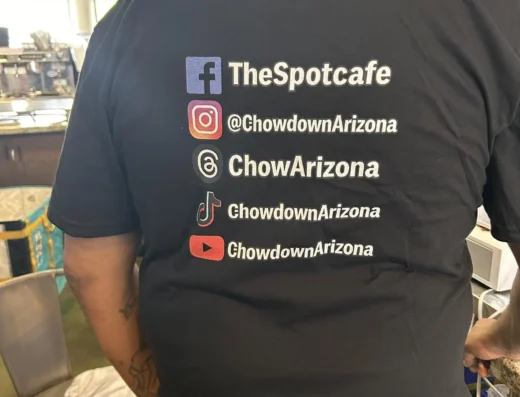 Back view of a person in a black T-shirt displaying social handles: TheSpotcafe on Facebook, ChowdownArizona on Instagram, TikTok, and YouTube.