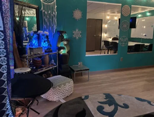 Teal-walled meditation room with a fountain sculpture, dreamcatchers, crystal decor, and blue fairy lights; a low table and soft poufs sit on a rug, with a doorway to a conference room in back.