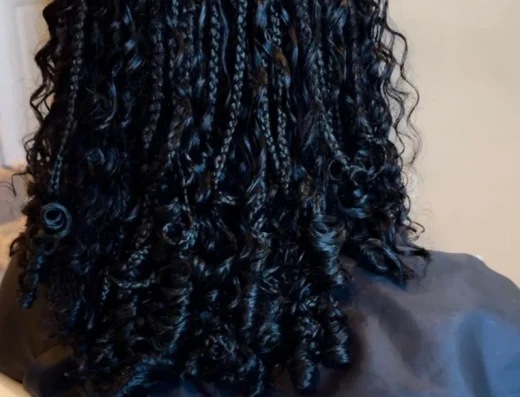 Back of person’s head with neatly braided cornrows and curly hair at ends, wearing a dark shirt in an indoor setting.