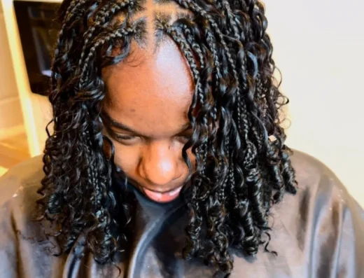 Person with neatly braided cornrows, parting down the center, long curly hair at the sides, wearing a dark cape in a salon.