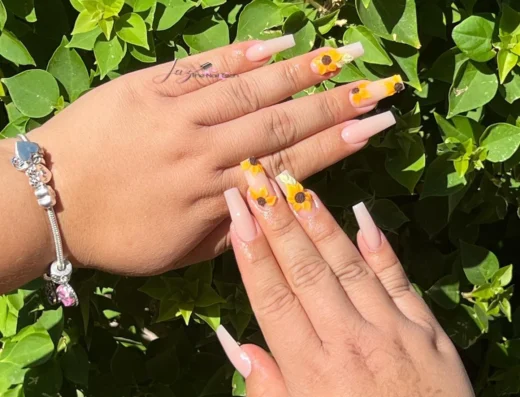 Hands showing nail art with long square nude nails and yellow daisy designs against green leafy background silver bracelet visible on wrist