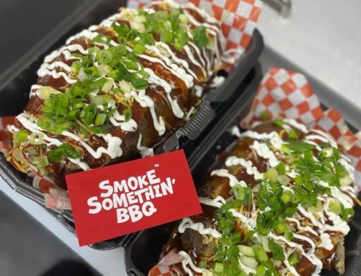 BBQ skewers glazed with sauce, sprinkled with chopped green onions, in a black takeout tray with red checker liner and a red 'Smoke Somethin' BBQ' card nearby.