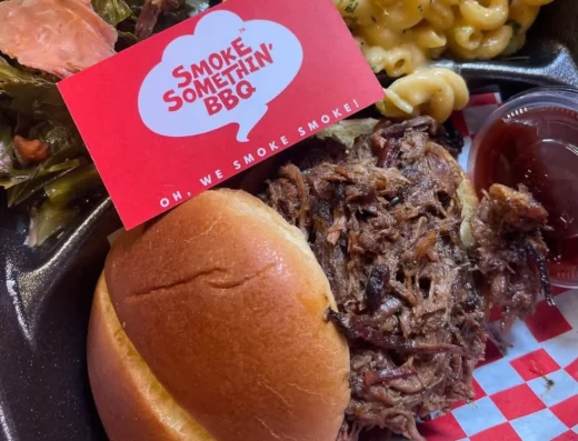 BBQ plate with a pulled pork sandwich, creamy macaroni and greens, and a side sauce; red Smoke Somethin' BBQ card on top of the dish.