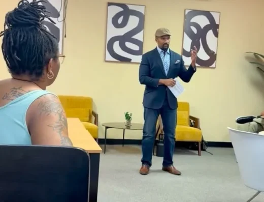 A man in a blue suit and brown hat speaks to an audience in a meeting room, holding notes.