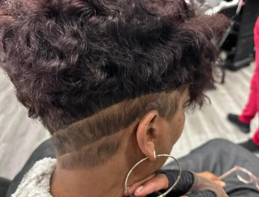 Person with a short, maroon-tinted, curly hairstyle and shaved sides in a hair salon, wearing a towel around the neck and a large hoop earring.