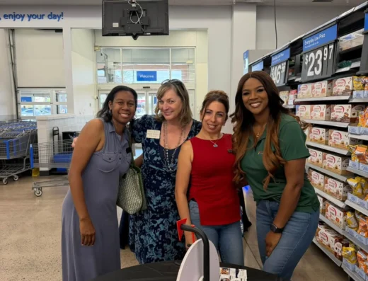 Four women smiling and posing together in a grocery store checkout area, with shelves of products visible in the background.