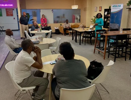 Conference room presentation with a woman in a teal dress speaking to a small audience seated at round tables.