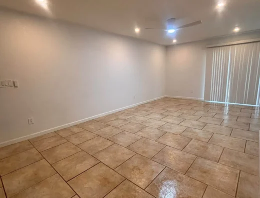 Empty living room with beige tile flooring, white walls, and a ceiling fan; sliding glass doors with vertical blinds on the right.