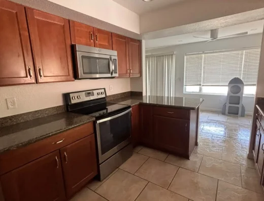 Modern kitchen with wood cabinets, stainless steel microwave and oven, granite counters, and tiled floor; open sightline to living area with blinds and ceiling fan
