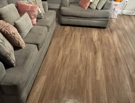 Living room scene with two gray couches and multiple pink and patterned throw pillows, wood-look flooring, and closed window blinds behind them.