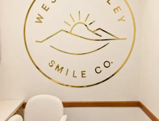 Gold circular logo of West Valley Smile Co with a sun and mountains on a light wall above a white desk and chair