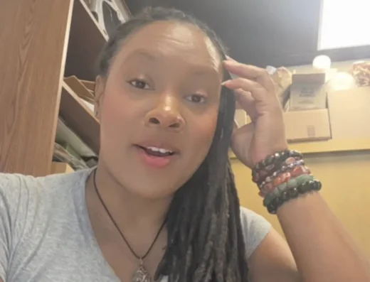 Smiling woman with long dreadlocks wearing a gray shirt, resting her hand on her head in an office space, wearing colorful beaded bracelets.