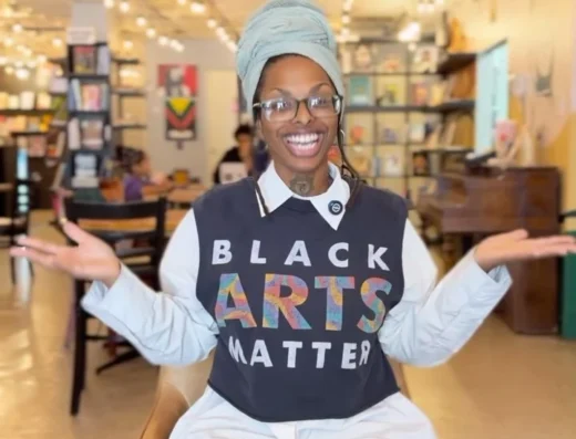 Smiling person with glasses and a blue head wrap sits in a bookstore, wearing a black 'Black Arts Matter' top over a white shirt; cheerful greeting text overlay reads 'Hello again from Grassroots Bookstore'
