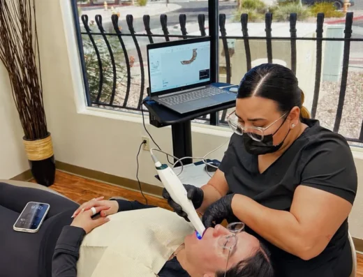 Dental professional in mask and gloves using a handheld tool on patient lying in a dental chair; laptop on a stand beside them.
