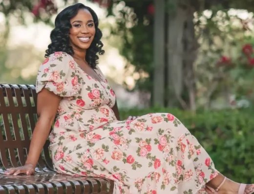 Pregnant woman in a floral dress sits on a park bench, smiling in sunlight garden setting.
