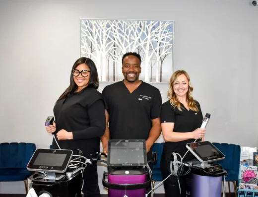 Three smiling staff members in black scrubs stand behind automated treatment machines in a modern clinic reception area.