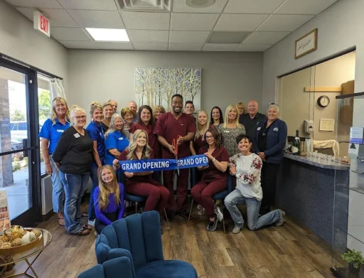 Group of about 20 people posing for a grand opening ribbon cutting event in a bright clinic like lobby several are in scrubs and name badges and two hold a blue ribbon that reads GRAND OPENING while the group smiles at the camera