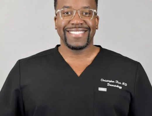 Smiling male dermatologist in black scrubs with clear glasses, against a light gray background.