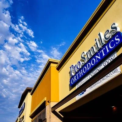 Exterior view of a yellow orthodontics clinic storefront with a sign reading 'ProSmiles Orthodontics' and 'Coming Soon' against a blue sky.