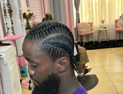 Man with neatly braided cornrows in a salon, shown in profile with pink decor in the background