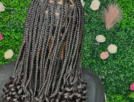 Back view of a person with tight, black box braids cascading down the back, ends curled, against a green hedge backdrop with pink and white roses and a pink neon sign nearby.