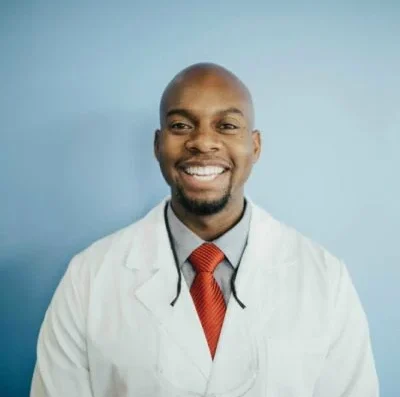 Smiling Black male doctor in a white coat and red tie, with a stethoscope around his neck, against a light blue background.