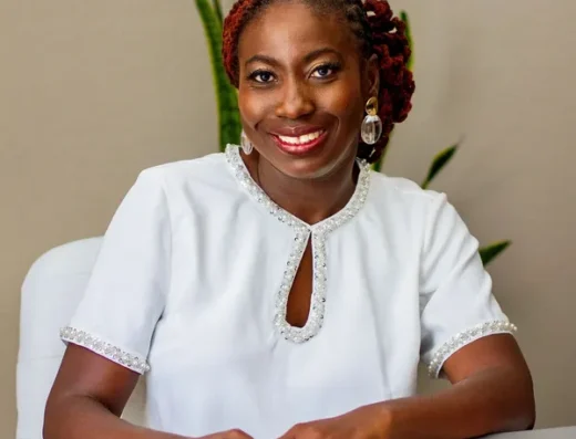 Smiling Black woman in a white embellished blouse sits at a desk with hands folded, plants in the background, in an office setting.