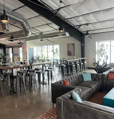 Industrial coworking cafe interior with exposed ceiling ducts, a long communal table with metal stools, and a gray sectional sofa with turquoise and orange cushions.