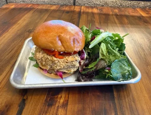 Crispy fried chicken sandwich on a glossy bun with tomato and greens on a metal tray beside a mixed greens salad on a wooden table.