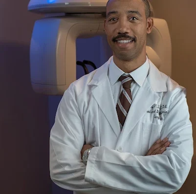 Radiologist in a white coat smiles with arms crossed, MRI machine visible in the background.